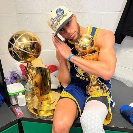Curry in a champions cap with the NBA trophy and Finals MVP trophy
