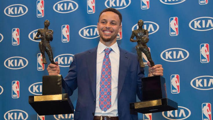 Curry in a suit standing between his two MVP trophies